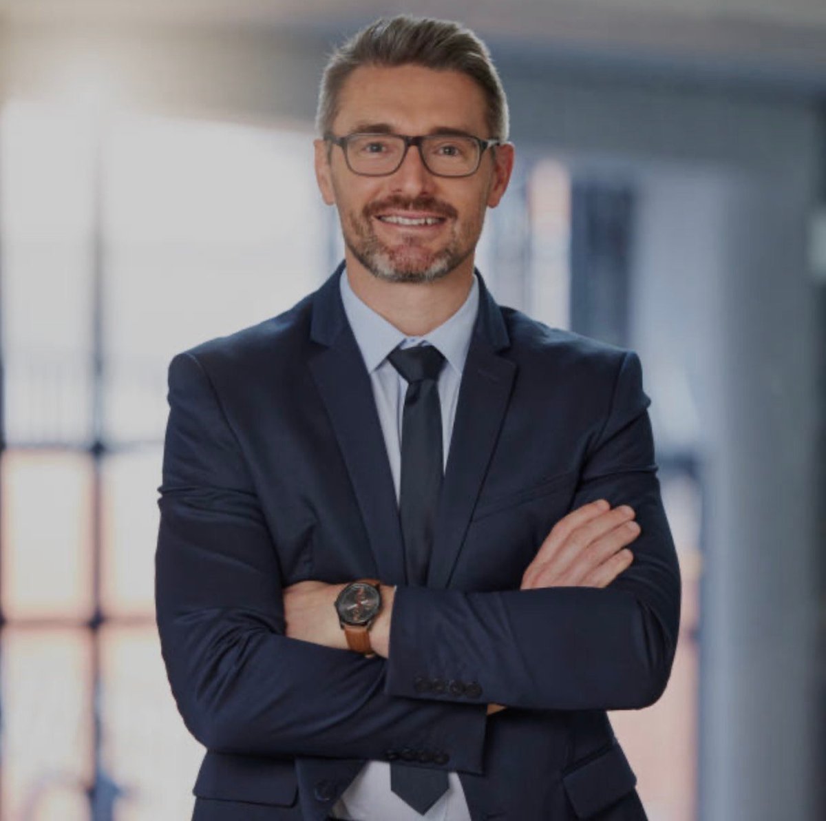 Professional headshot of a man in business attire with crossed arms, wearing glasses and a dark suit in an office setting