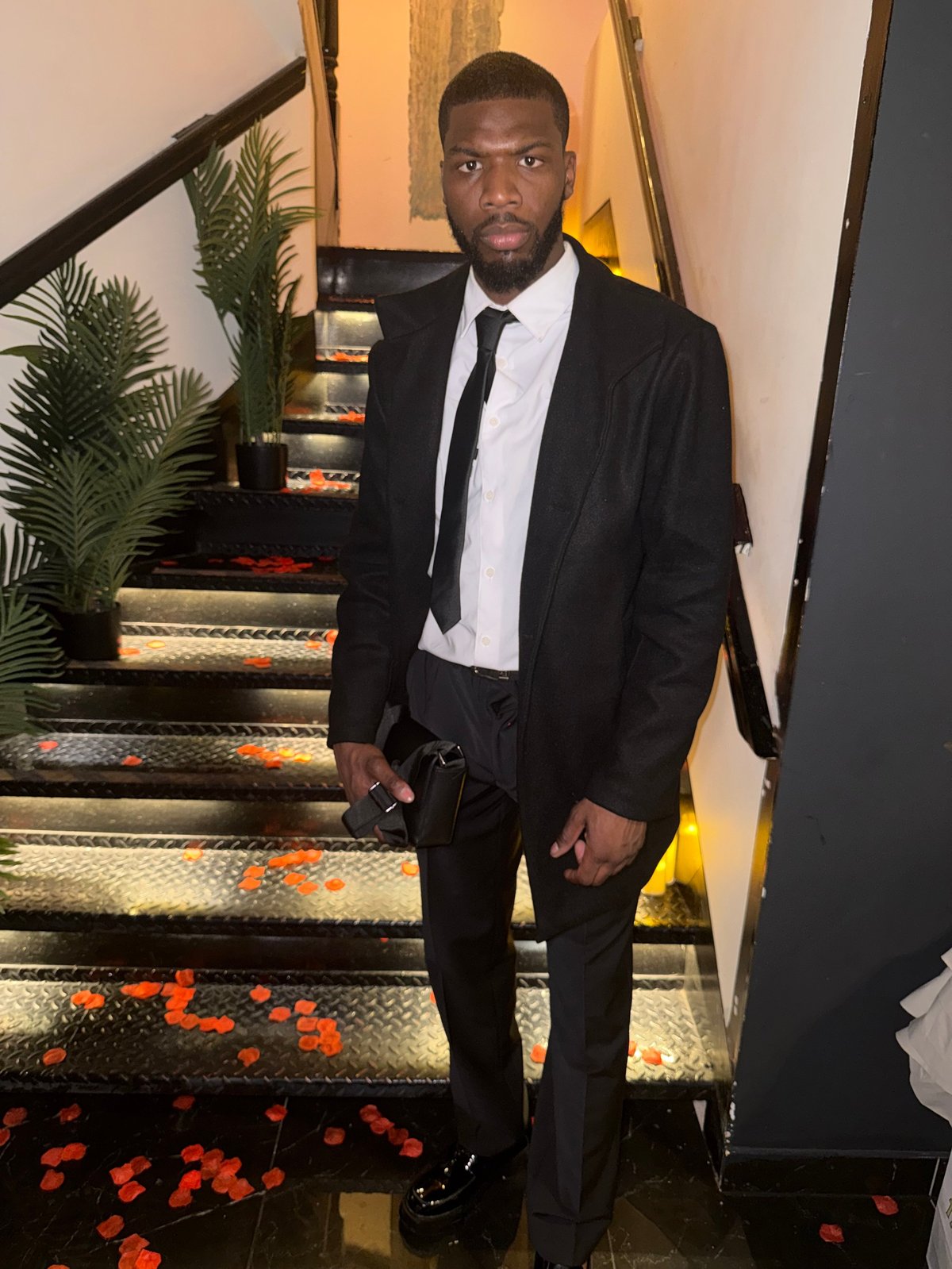 Man in formal black suit and white dress shirt standing on decorative staircase with potted plant in background